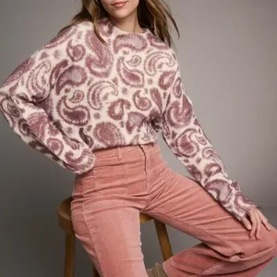 Mulher com casaco de malha padrão creme e rosa e calças rosa de veludo cotelê sentada num banco de madeira