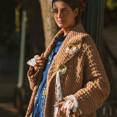 Mulher com casaco castanho de malha grossa e camisa azul bordada, apoia-se num poste verde em ambiente exterior.