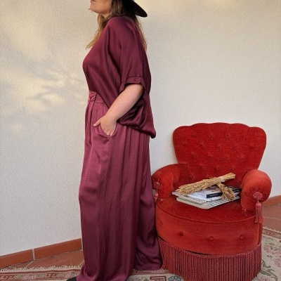 Mulher de perfil com vestido vermelho vinho e chapéu preto junto a poltrona vermelha com livros