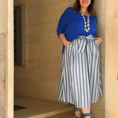 Mulher com camisola azul e saia às riscas brancas e cinzentas com laço, colar branco, sandálias verdes com cordão, encostada a parede de pedra.