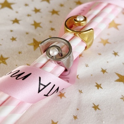 Dois anéis metálicos com pérolas em palhinhas cor-de-rosa e branca rodeados por fita rosa com texto