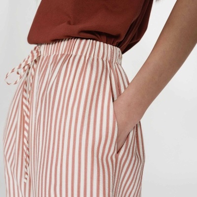 Calças riscas verticais brancas e terra com cintura elástica e cordão, com mão no bolso, vestidas com t-shirt castanha