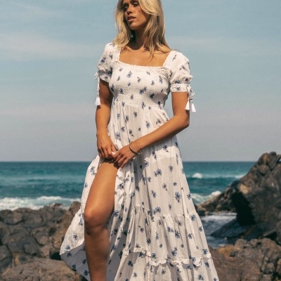 Mulher com vestido longo branco floral em praia rochosa.