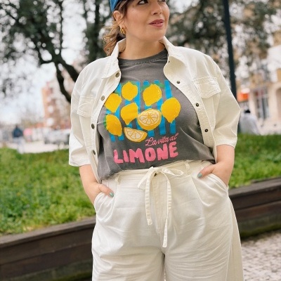 Mulher com t-shirt cinzenta estampada de limões, calças creme, camisa creme aberta e boné azul