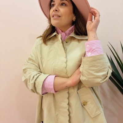 Mulher vestindo casaco bege e chapéu rosa, com camisa rosa às riscas e jeans azuis, em fundo rosa com planta.