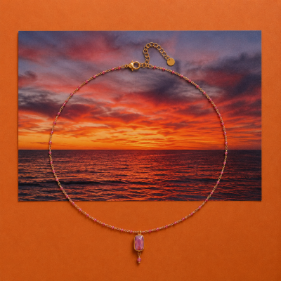 Colar dourado com contas rosadas e pendente roxo sobre fundo laranja e fotografia de pôr do sol no mar.