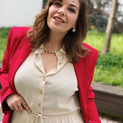 Mulher com camisa creme, casaco vermelho e acessórios dourados num ambiente exterior com relva.
