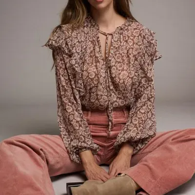 Mulher sentada a usar blusa floral com folhos, calças rosa de veludo cotelê e botas castanhas.