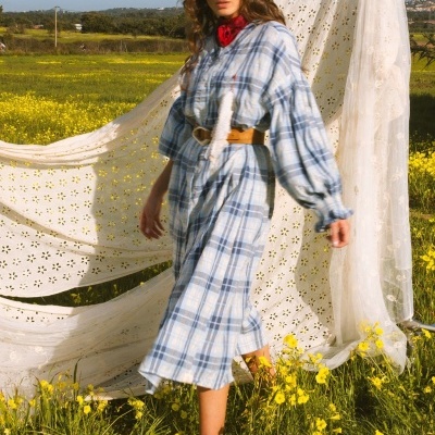 Mulher a usar vestido xadrez azul claro e branco, cinto castanho, lenço vermelho e óculos de sol, em campo florido com tecido branco decorativo ao fundo.