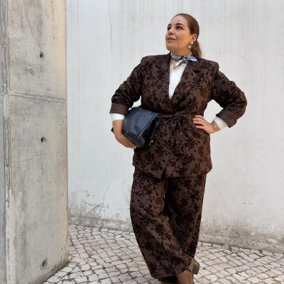Mulher com fato e botas castanhas, segurando bolsa preta, com lenço ao pescoço, parede branca e chão em mosaico claro.