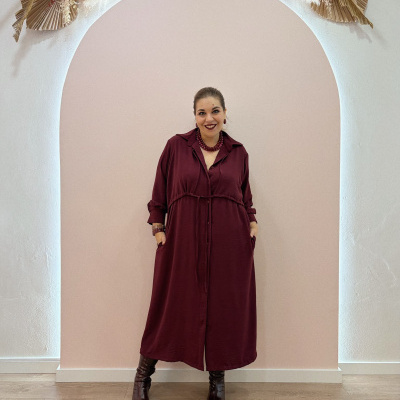 Mulher com vestido bordô comprido em frente a parede cor de rosa claro com decoração de flores secas acima.