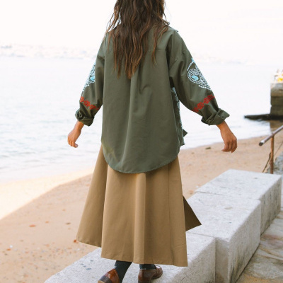 Pessoa de costas a usar blusa verde com estampados brancos e vermelhos, saia bege e sapatos castanhos junto à praia