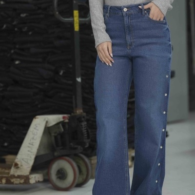 Calças de ganga azul com botões laterais e blusão cinzento usado por mulher em ambiente industrial