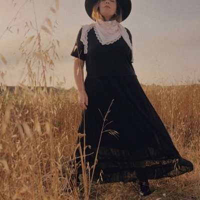 Mulher com vestido preto, lenço com renda branca e chapéu preto em campo seco ao pôr do sol