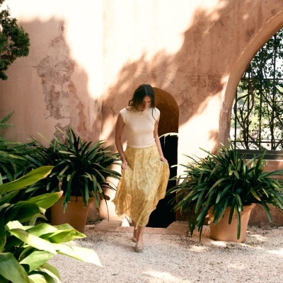 Mulher com vestido amarelo e top branco num jardim com plantas verdes em vasos.