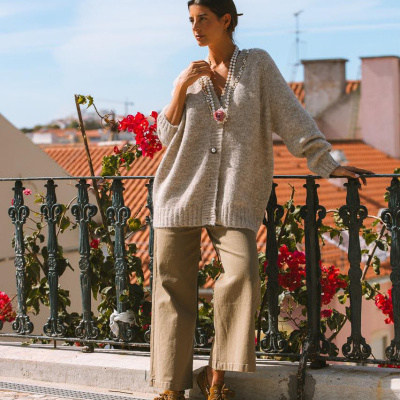 Mulher com cardigan cinza e calças bege numa varanda com corrimão de ferro e flores vermelhas