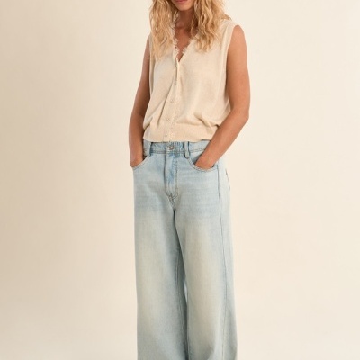 Mulher vestindo colete bege de malha com botões e calças jeans largas azul claro, em fundo neutro bege claro.