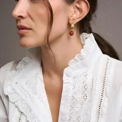 Mulher com brinco dourado de bola vermelha e camisa branca bordada com babados.