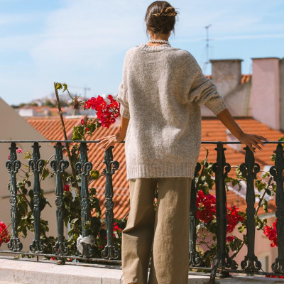 Pessoa de costas com camisola cinzenta, calças largas castanhas e sapatilhas castanhas numa varanda com flores vermelhas e casas ao fundo.