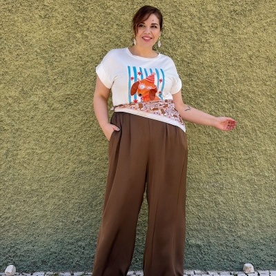 Mulher com t-shirt branca com estampa de cão, calças largas castanhas e lenço castanho posando em parede verde texturada.