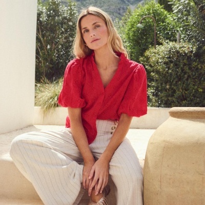 Mulher com blusa vermelha e calças largas bege num cenário exterior com plantas e vaso cerâmico.