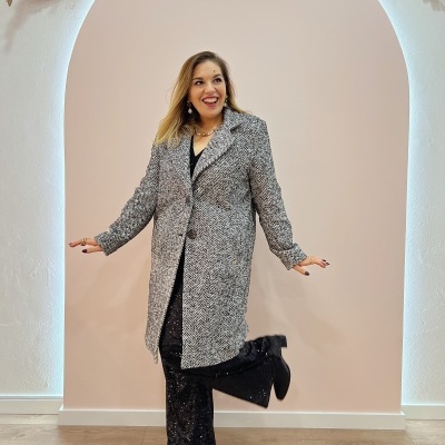 Mulher com casaco comprido cinza e branco, calças pretas brilhantes e botas pretas em interior com arco decorado.