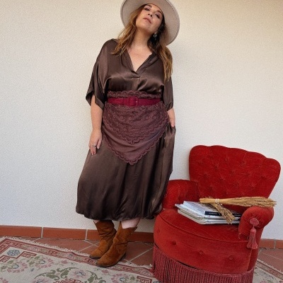 Mulher com vestido castanho, cinto vermelho e chapéu bege ao lado de poltrona vermelha vintage