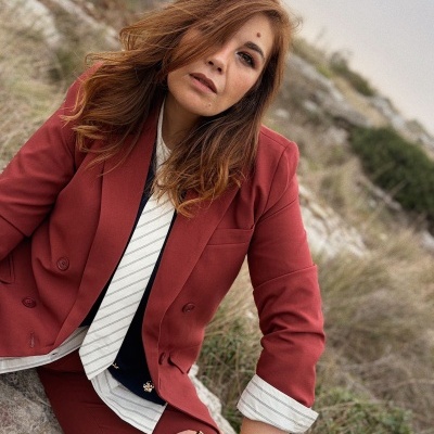 Mulher com casaco vermelho vinho e camisa branca às riscas a sentar em ambiente natural