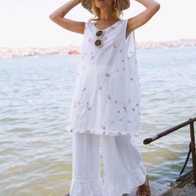 Mulher com vestido branco bordado, calças brancas largas, chapéu de palha e sandálias junto ao mar