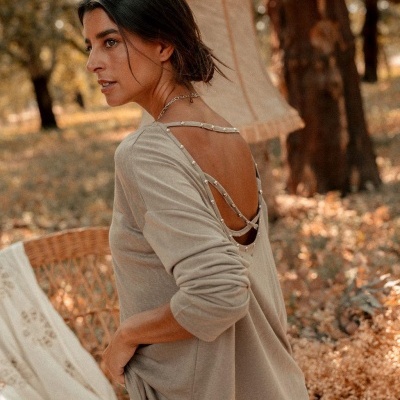 Mulher com camisola bege com decote em V nas costas e detalhes de tachas prateadas num ambiente natural