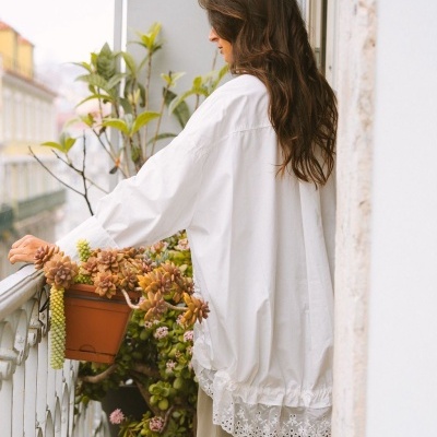 Blusa feminina branca com barra bordada e mangas compridas vista de costas em varanda com plantas