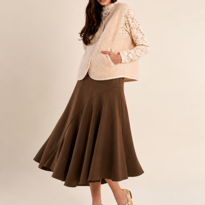 Mulher com blusa de renda creme, colete creme, saia castanha e sapatos dourados, fundo creme.