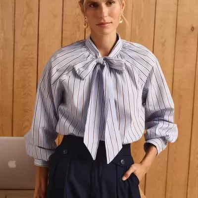 Mulher com camisa de riscas azul clara com laço e calças azul-escuras em ambiente de madeira