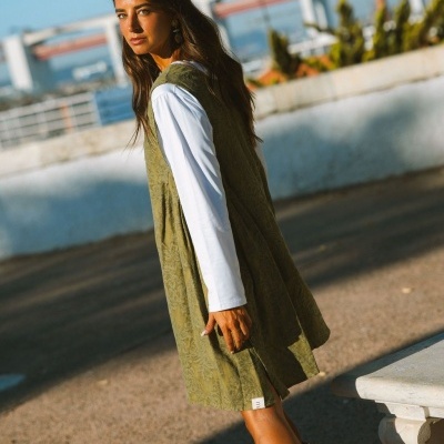 Mulher com vestido verde de padrão floral, camisa branca e botas castanhas em área urbana ao sol