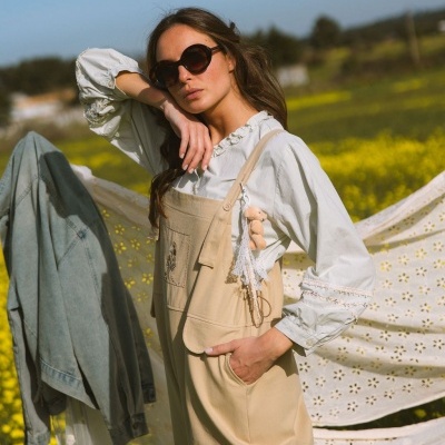 Mulher com óculos castanhos, blusa branca rendada e macacão bege em campo de flores amarelas