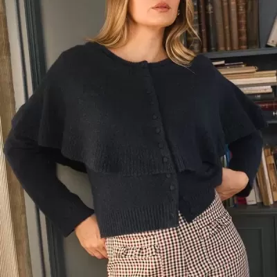 Mulher com casaco azul escuro de malha e calças de quadrícula em ambiente interior com estante de livros