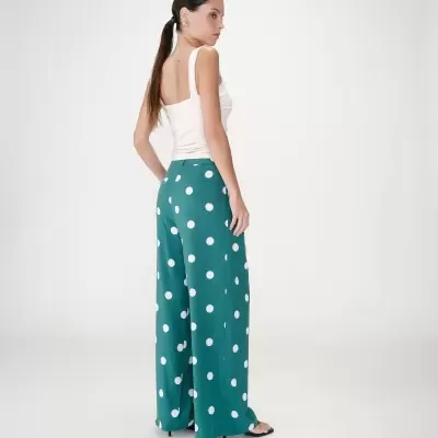 Mulher de costas com calças largas verdes com bolinhas brancas e top branco, em fundo branco.