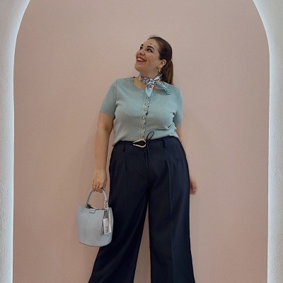 Mulher com camisola azul clara e calças pretas, sandálias castanhas, lenço azul no pescoço e mala azul clara, parede rosa ao fundo.