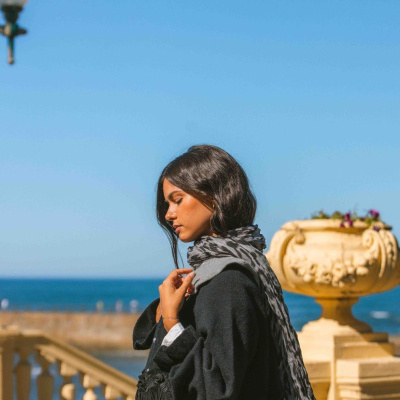 Mulher com cachecol cinza e casaco preto ao ar livre junto ao mar