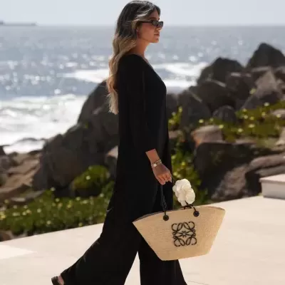 Mulher de vestido preto com mala de palha junto ao mar