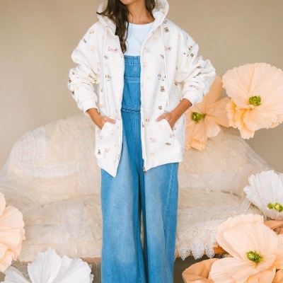 Mulher com casaco branco com padrão de desenhos, macacão de ganga, flores de papel e sofá rendado