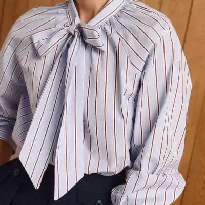 Camisa masculina azul clara às riscas com laço na gola e calças pretas, parede de madeira ao fundo