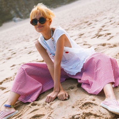 Pessoa com calças xadrez vermelho e branco, blusa branca, óculos de sol grandes e sapatilhas cor-de-rosa sentada na areia