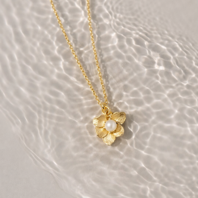Colar dourado com pendente de flor e pérola em fundo com reflexos de água