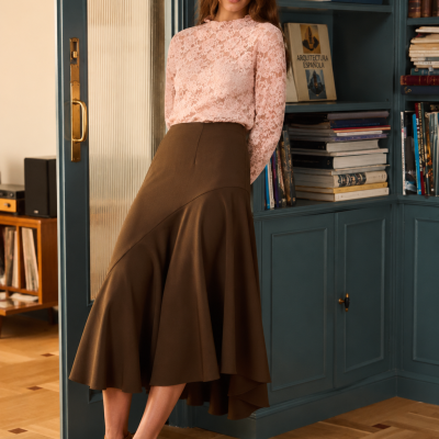 Mulher vestida com blusa rosa de renda e saia castanha, com sapatos castanhos, em interior com estante azul e livros.