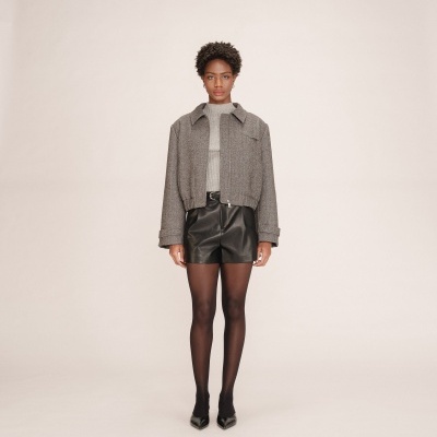 Pessoa com casaco cinzento, calções pretos, collants e sapatos pretos em fundo bege.