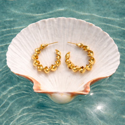 Brincos argola dourados ondulados numa concha branca sobre água azul
