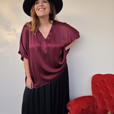 Mulher com blusa vinho acetinada, saia preta e chapéu preto em ambiente interior junto a cadeira vermelha