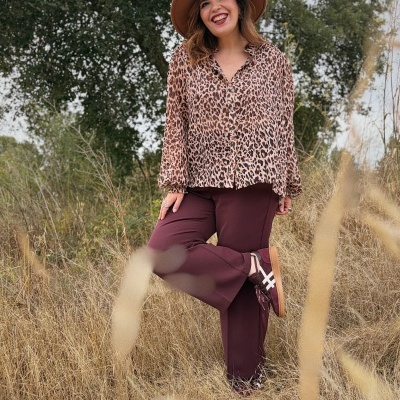 Mulher com blusa animal print, calças castanhas e chapéu castanho em ambiente natural
