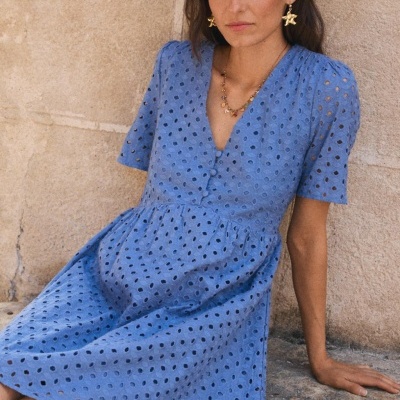 Vestido azul com recortes circulares e botões forrados, modelo a usar por mulher sentada junto a parede.